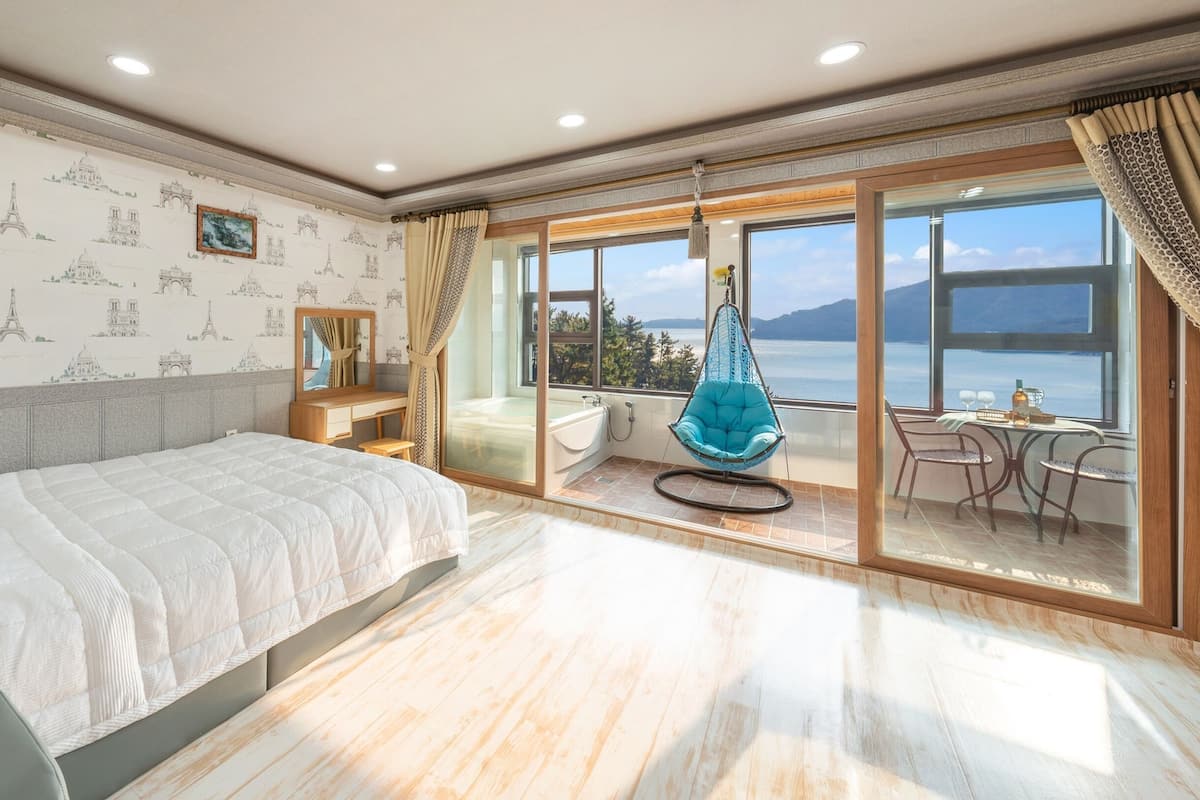 A spacious bedroom with a large bed, a sitting area with a hanging chair, and a balcony with a view of the sea.