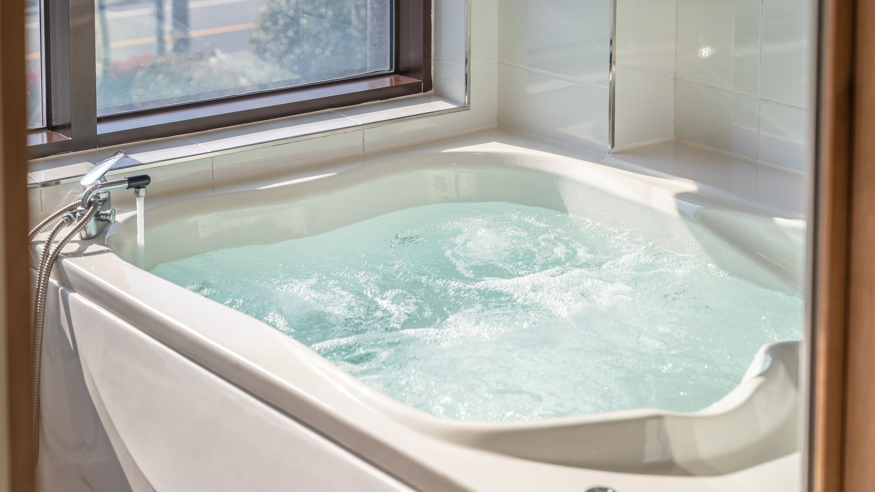 Une baignoire remplie d’eau, une fenêtre au-dessus et des murs recouverts de carreaux blancs.