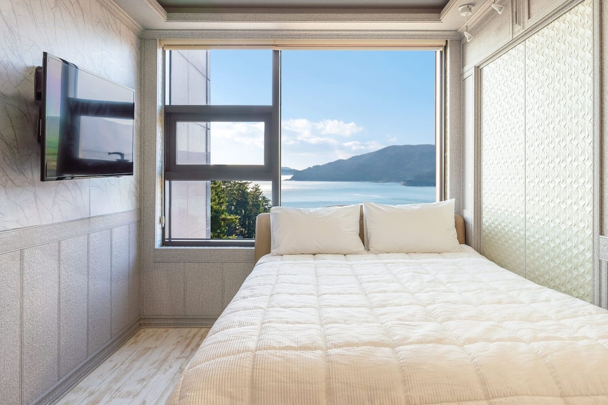 A bedroom with a large window offering a view of the sea and hills.