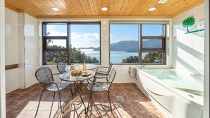 Una habitación con techo de madera, ventanas amplias con vista a un lago y montañas, una mesa redonda con sillas y un jacuzzi.