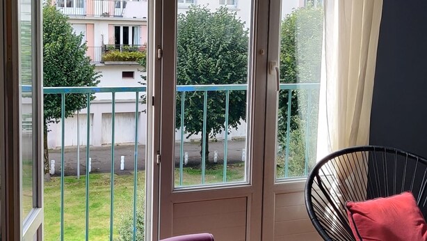 Et værelse med en stor skydedør, der fører til en balkon, en lilla stol, en sort metalstol med rød pude og et ternet trægulv.