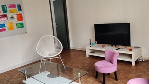 En stue med et glasbord, en hvid stol, en pink stol, et fjernsyn på et hvidt skab og et farverigt abstrakt maleri på væggen.