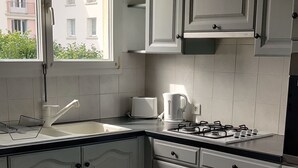Et køkken med hvide skabe, en gas komfur, en kaffemaskine og et vindue med udsigt til bygninger.