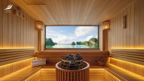 Una sala de sauna con bancos de madera, una hoguera en el centro y un ventanal con vistas a un lago y montañas.