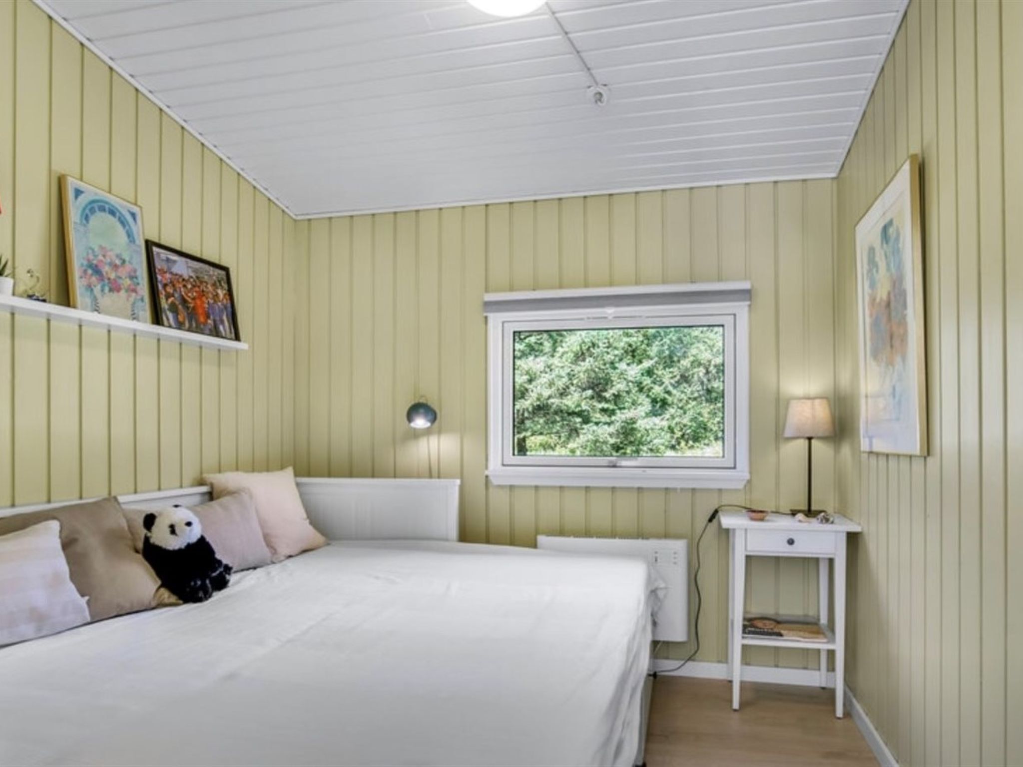 A bedroom with a bed, a nightstand, a window with a view of trees, and wall-mounted shelves with pictures.