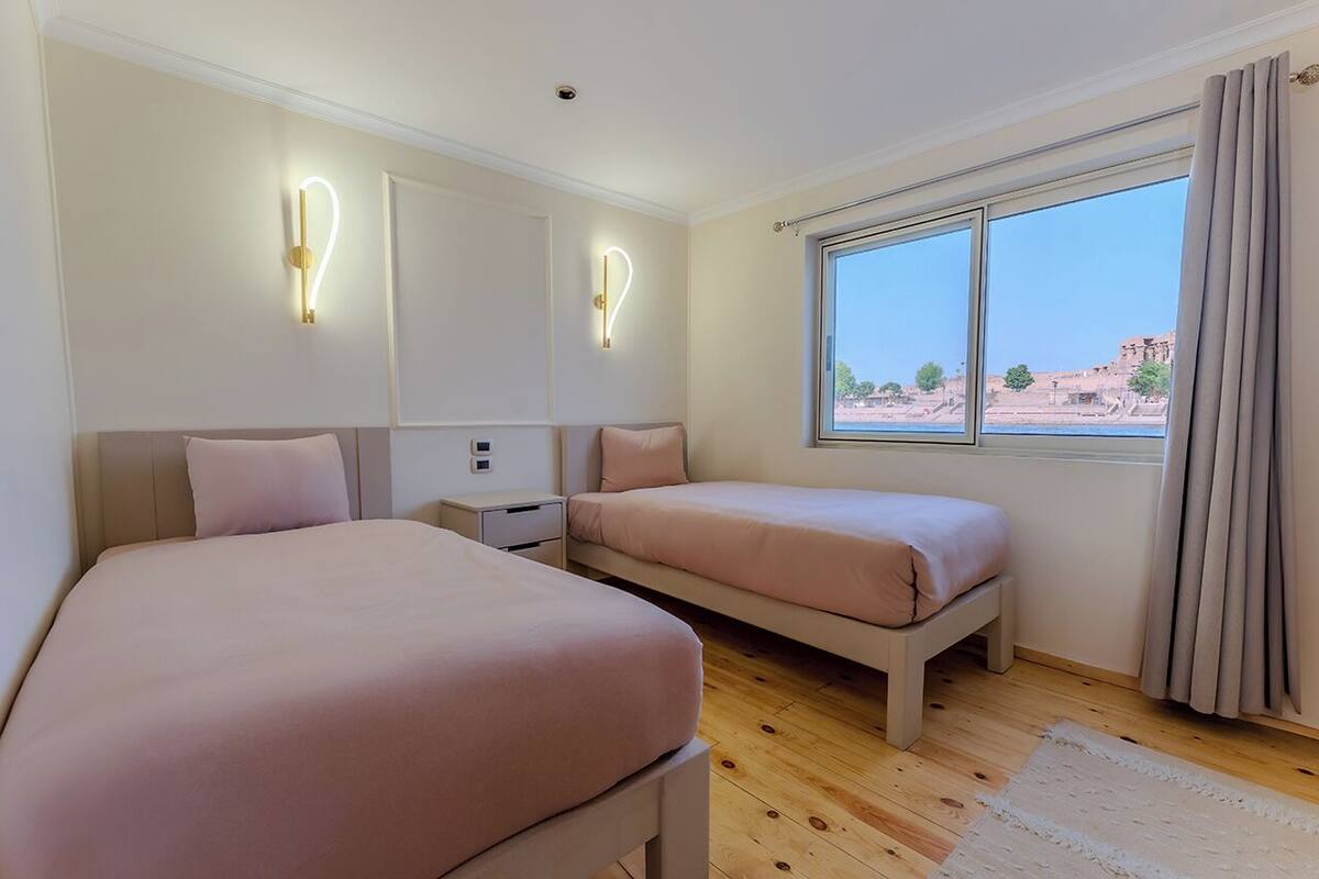Un dormitorio con dos camas, una mesita de noche, una lámpara de pared y una ventana con cortinas.