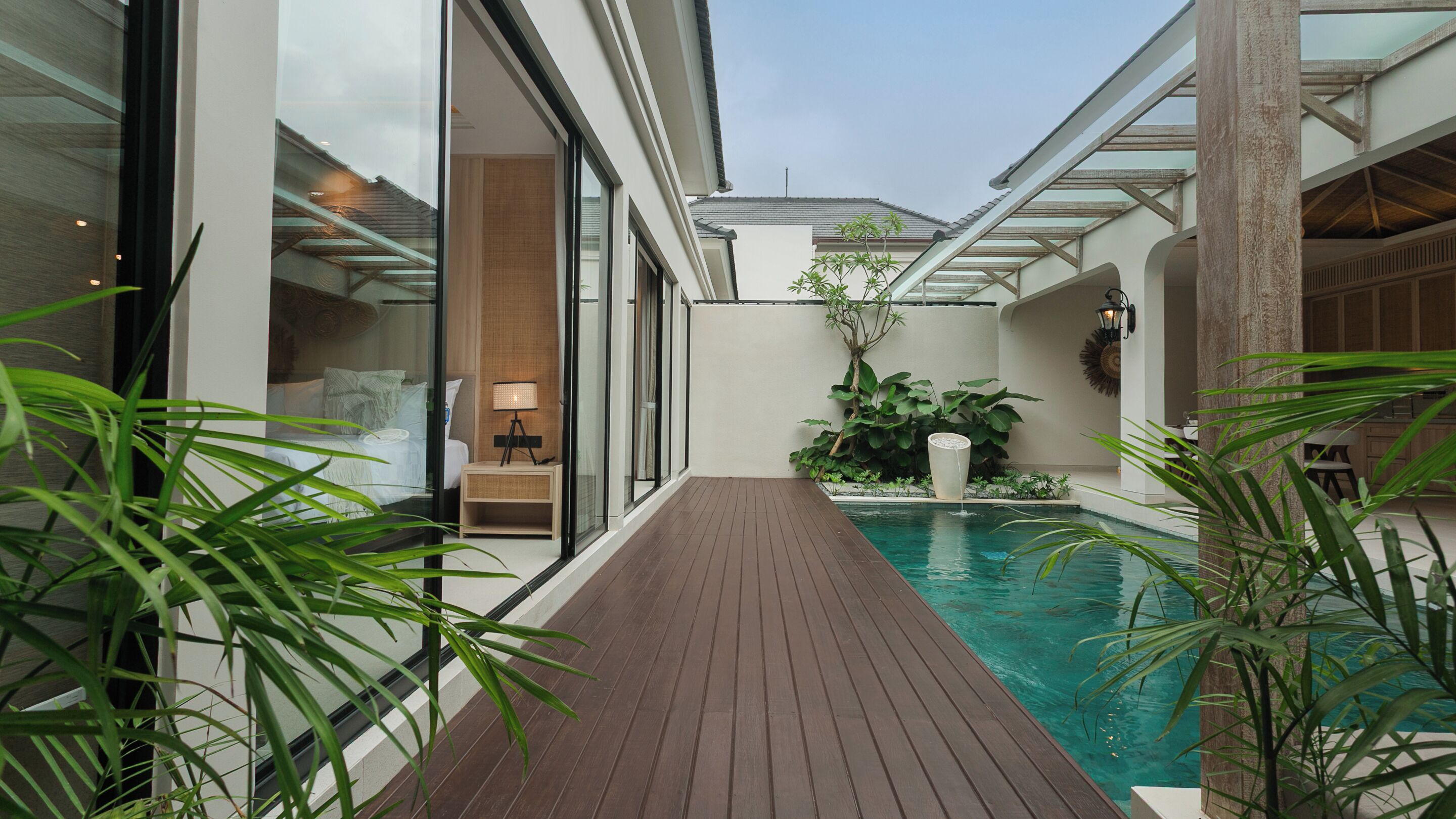 A modern pool area with a wooden deck, glass walls, and a small seating area with a lamp.