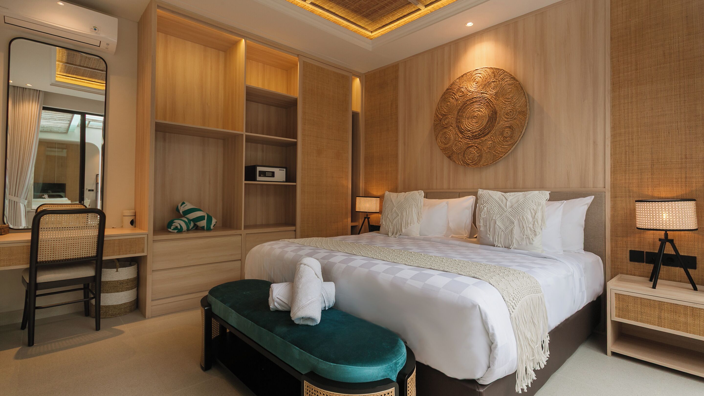 A hotel room with a large bed, a desk with a chair, a bench, and a wall-mounted round decorative piece.
