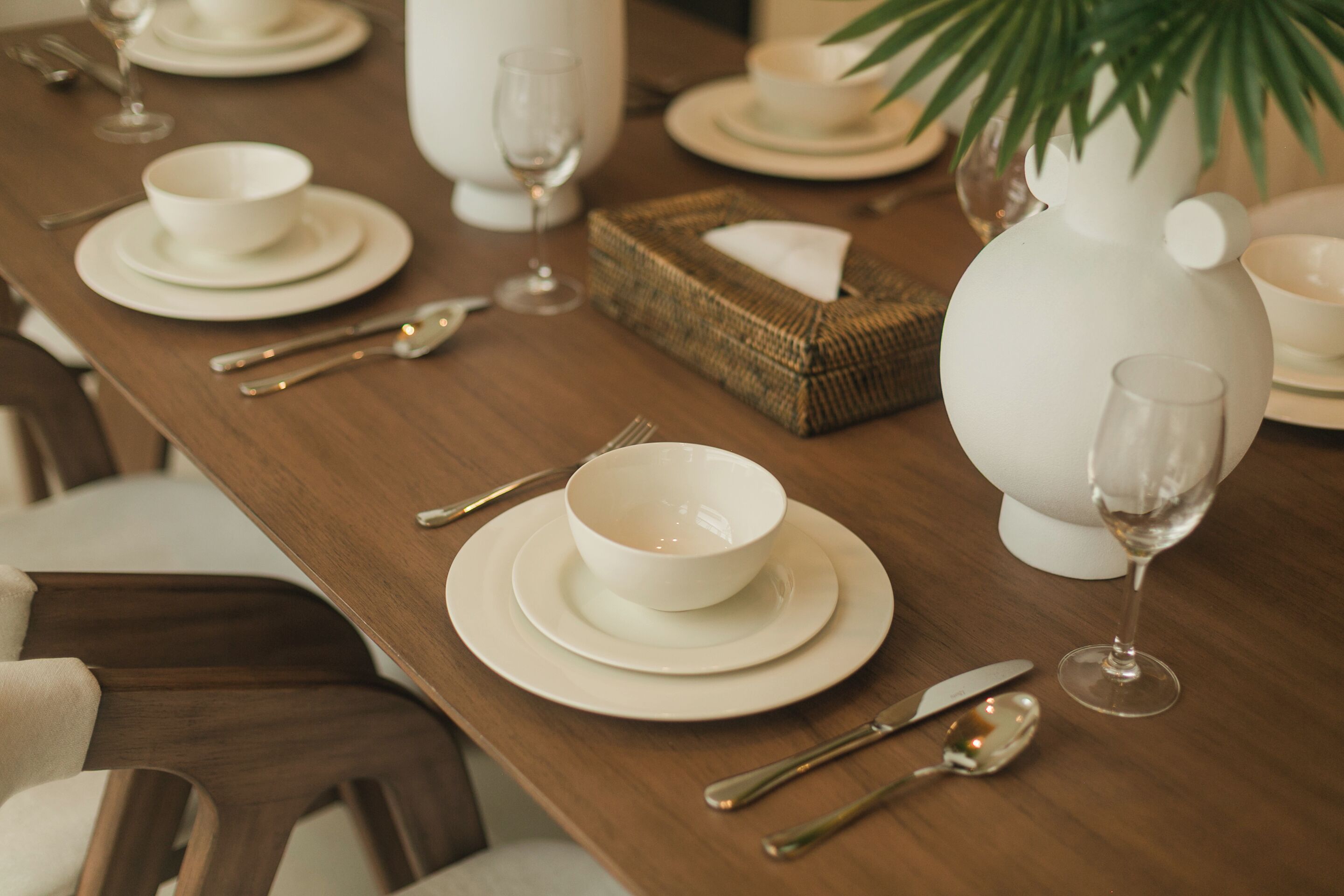 Mesa de comedor dispuesta con platos, cuencos y cubiertos blancos, incluyendo un jarrón y una copa.
