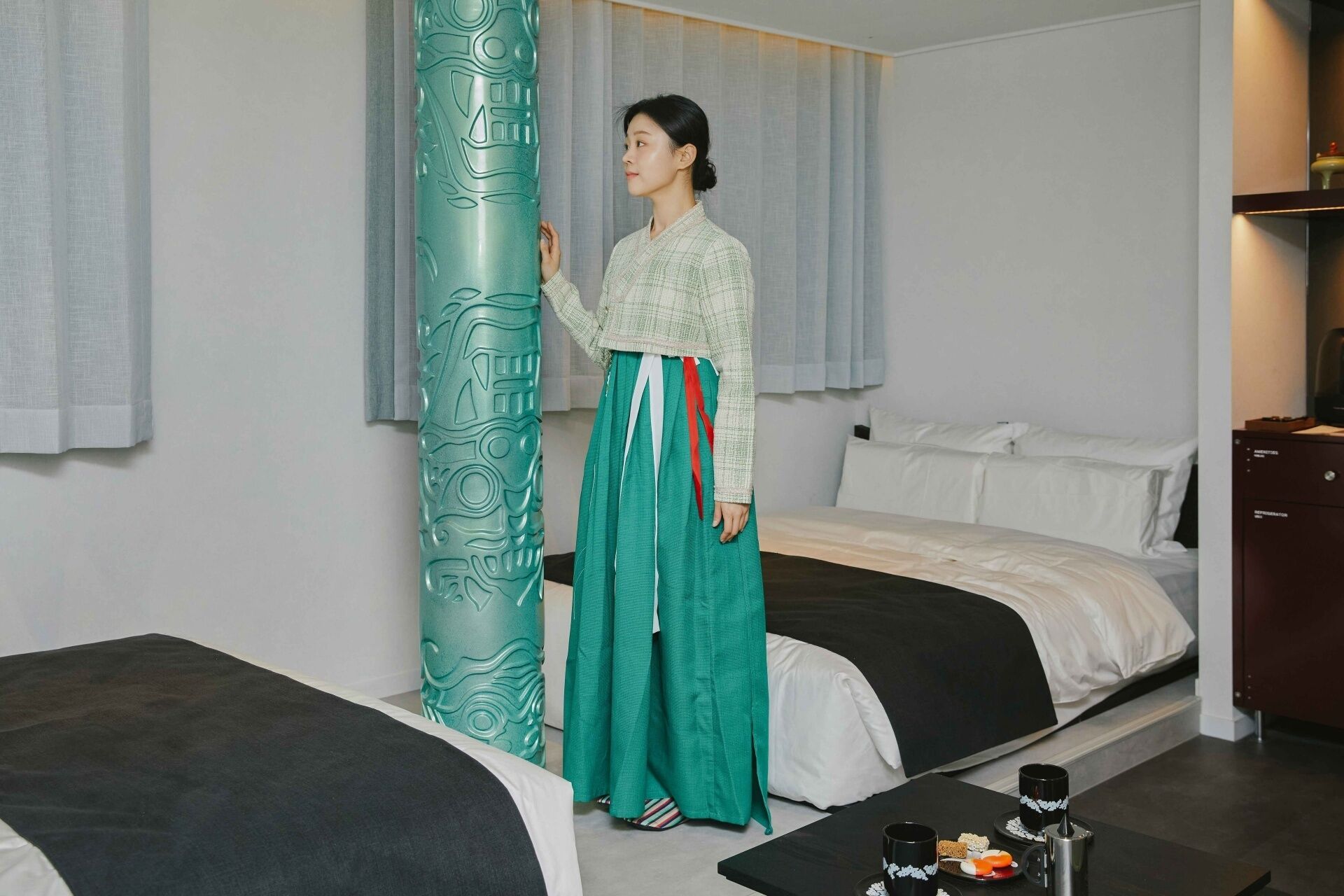 A hotel room with two beds, a large teal decorative pillar, and a small table with a coffee pot and cups.