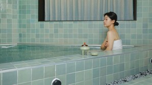 Una persona en una bañera con una toalla sobre el hombro, un recipiente pequeño en el borde y una pared con azulejos y una cortina.