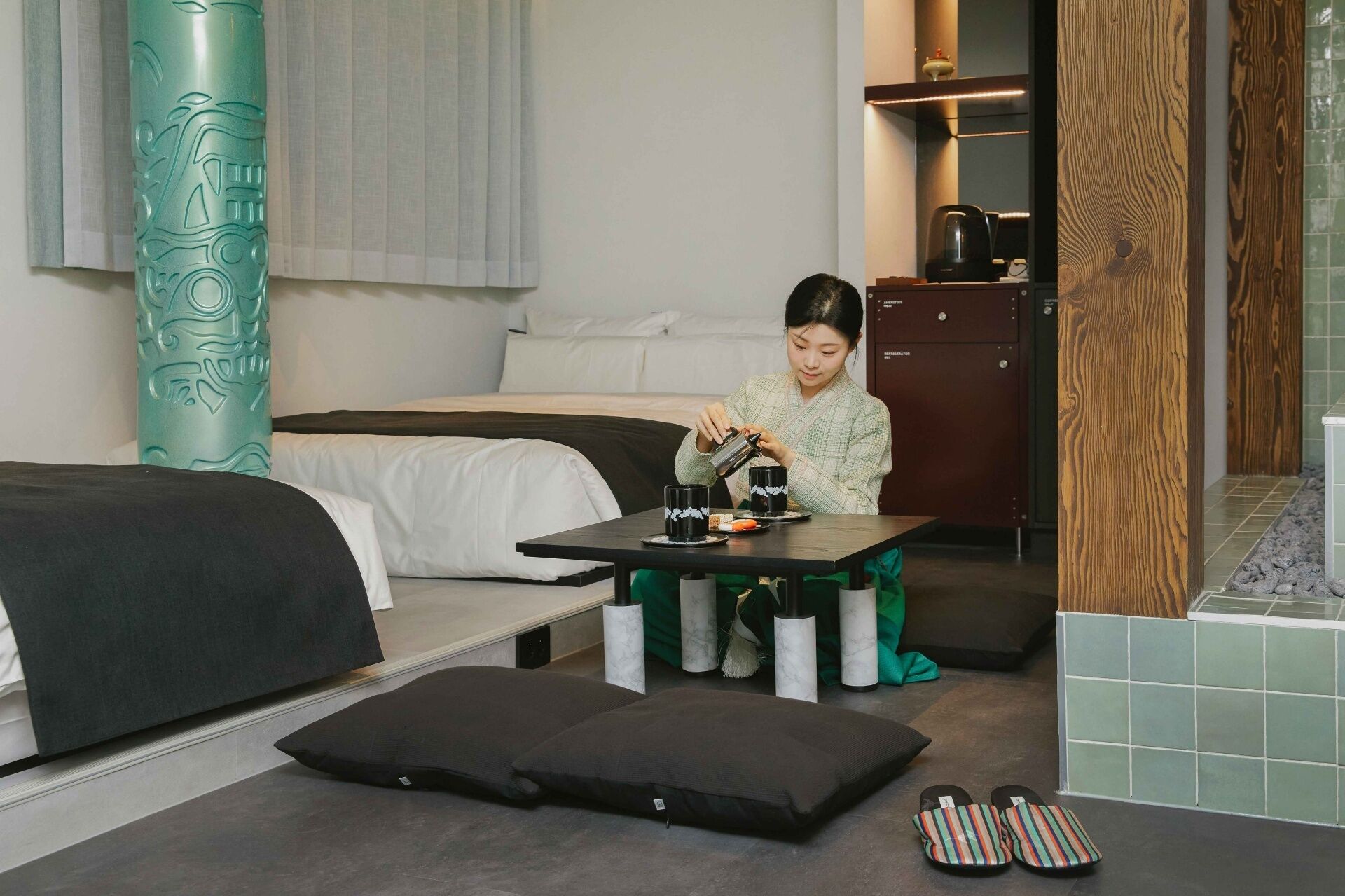 A hotel room with a bed, a small table with two cups, a kettle, and slippers on the floor.