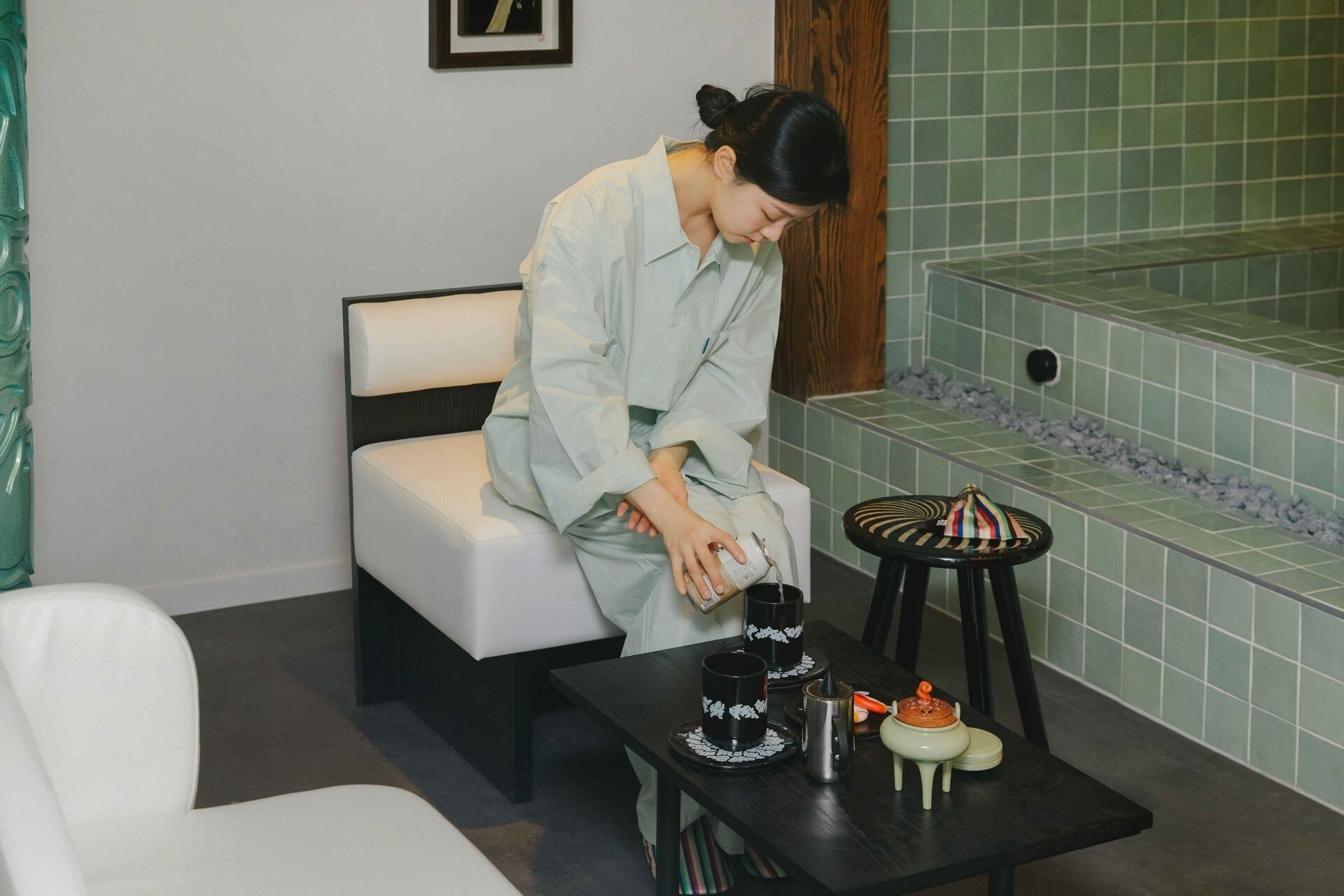 A person sitting on a white sofa in a room with a green tiled wall and a small table with a teapot and cups.