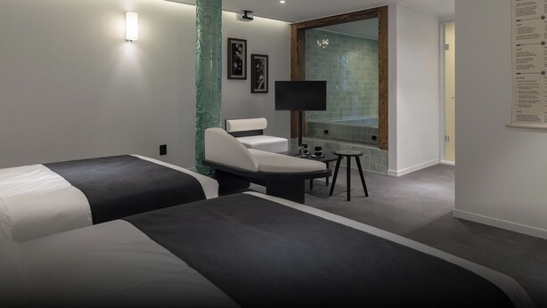Habitación de hotel con dos camas, zona de estar, una mesita y baño con cabina de ducha de vidrio.