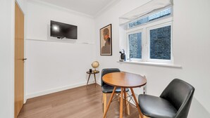 Una habitación con piso de madera, una mesa redonda con sillas, un televisor montado en la pared, un cuadro enmarcado y una ventana con vista a árboles.