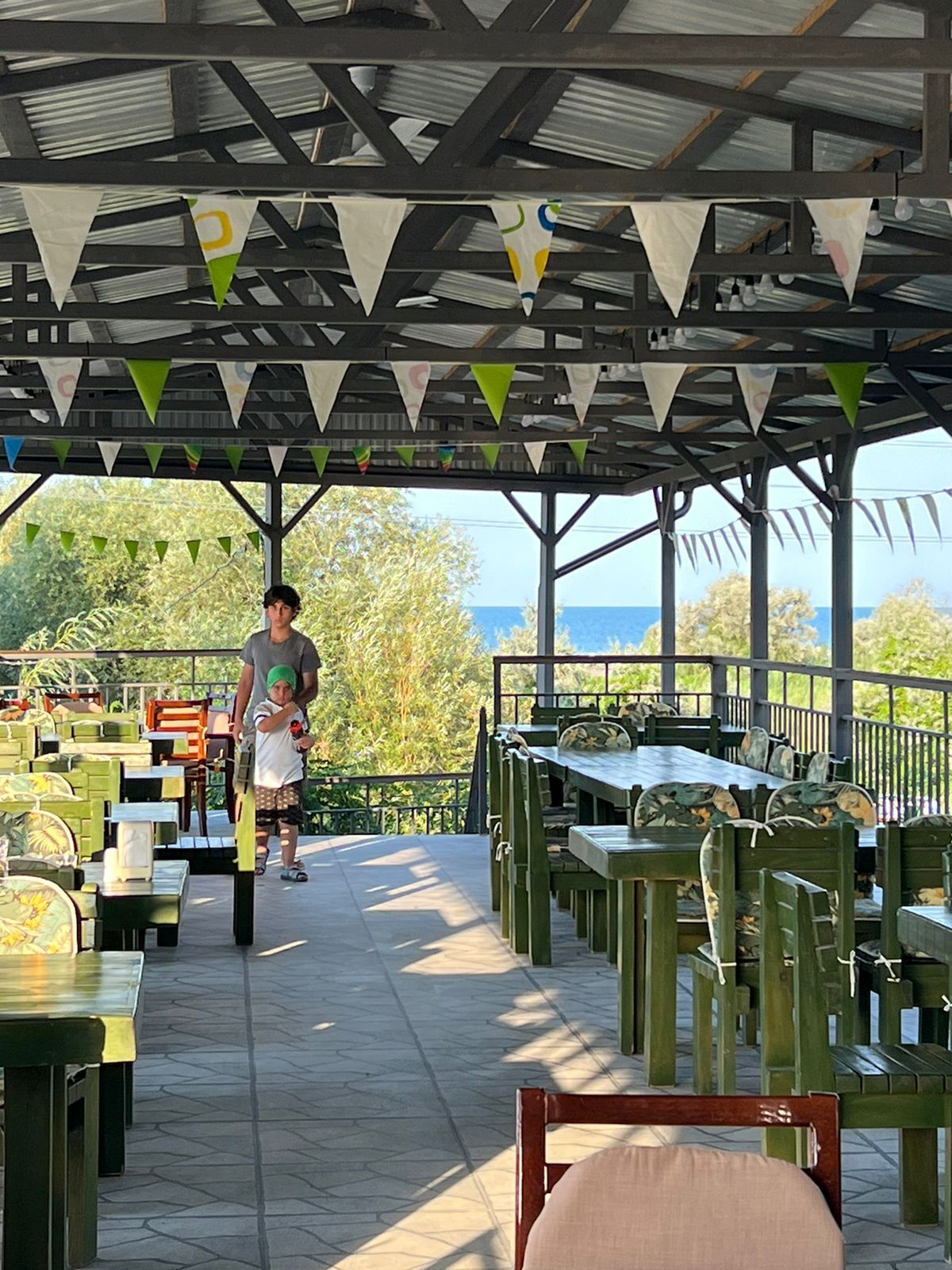 An outdoor dining area with tables and chairs under a covered roof, surrounded by greenery and a view of the sea.