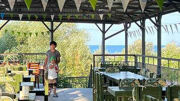 An outdoor dining area with tables and chairs under a covered roof, surrounded by greenery and a view of the sea.