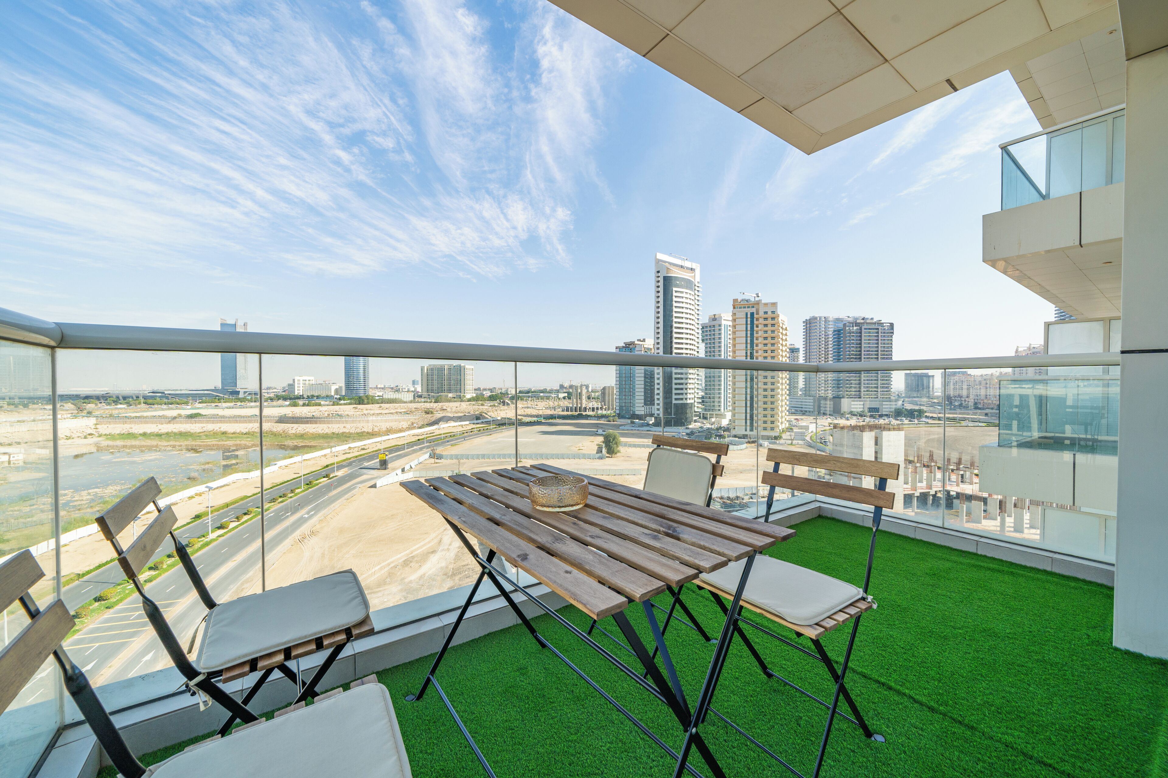 Un balcón con mesa y sillas de madera, vistas a un paisaje urbano y césped artificial.
