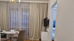 Ein modernes Wohnzimmer mit Essbereich, Holzboden, einem an der Wand montierten Fernseher und einem großen Fenster mit Vorhängen.