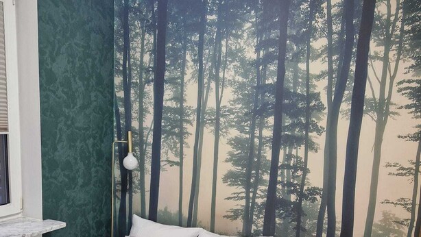 Ein Schlafzimmer mit Bett, Nachttisch, Lampe und einer Wandmalerei eines Waldes.