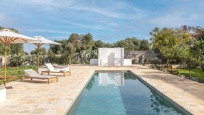 Une piscine rectangulaire avec une terrasse en pierre, deux chaises longues et une pergola dotée de parasols.