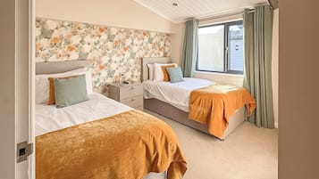 Une chambre avec deux lits, une tête de lit à motifs floraux, une fenêtre avec des rideaux et un plafond doté d’un éclairage encastré.