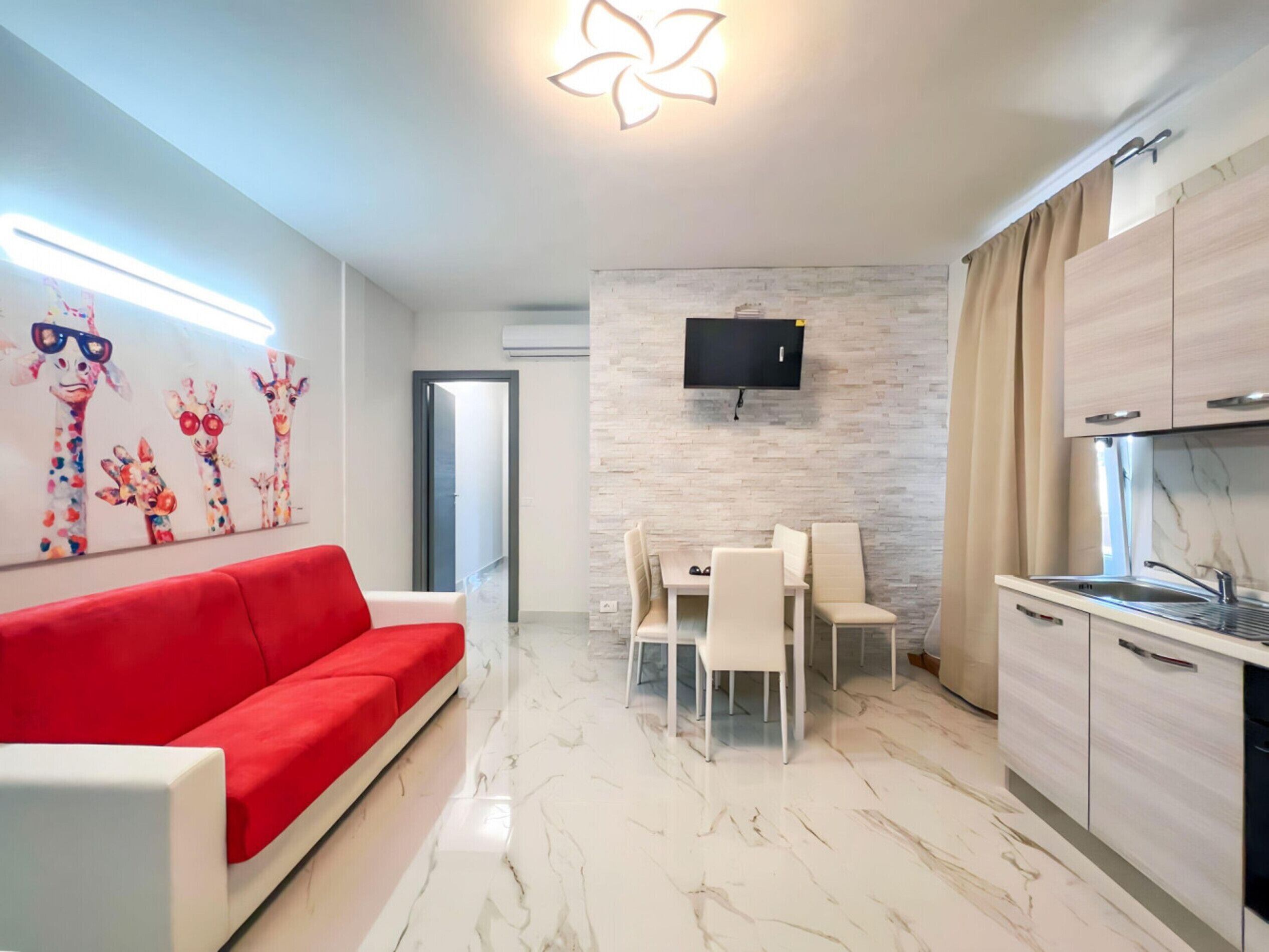 Uma sala de estar moderna com um sofá vermelho, uma área de jantar com mesa e cadeiras, uma cozinha com uma TV fixada na parede e um lustre.