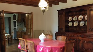 Una zona de menjador amb una taula de fusta coberta per un mantell rosa, envoltada de cadires de vimet. Hi ha una làmpada d'oli penjant del sostre, i un armari de fusta amb plats decoratius es pot veure al fons.