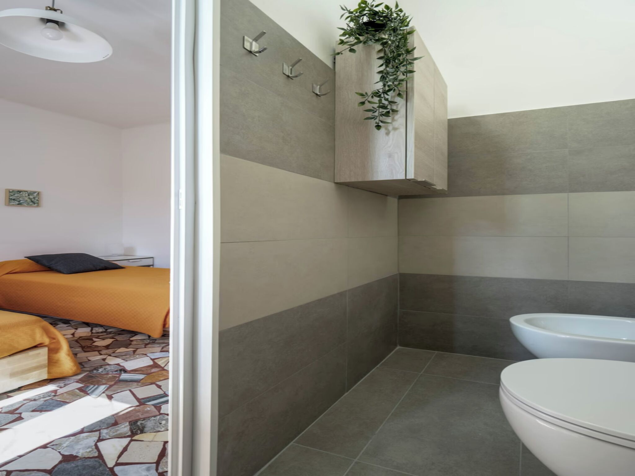 A modern bathroom with a toilet, a shower area, and a sink. There is a bed with an orange bedspread in the adjacent room.
