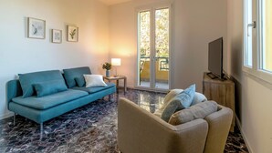 A living room with a blue sofa, a beige armchair, and a TV on a wooden stand. There is a small table with a lamp and a plant. The room has a large window with a view of trees. - Room-charming Guest Room in Frassino wit (Castel San Pietro Terme)