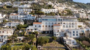 Une ville pittoresque, construite sur une colline et caractérisée par ses bâtiments blancs, dont certains sont dotés de balcons et de fenêtres en arc de cercle, le tout entouré de verdure et d’arbres.