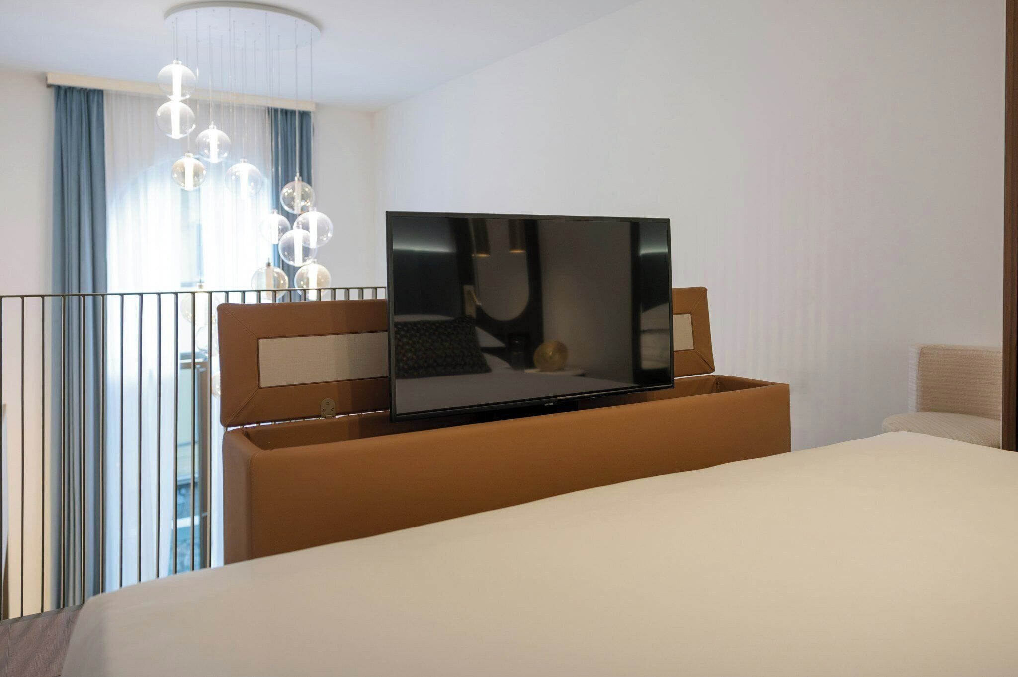 A modern hotel room with a large bed, a flat-screen TV mounted on a wooden headboard, a balcony with a railing, and a chandelier.