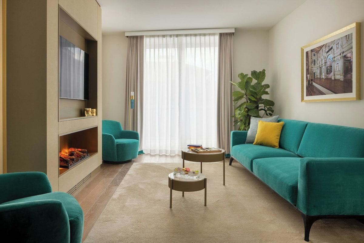 A modern living room with teal sofas, a fireplace, a flat-screen TV, and a large window with sheer curtains.