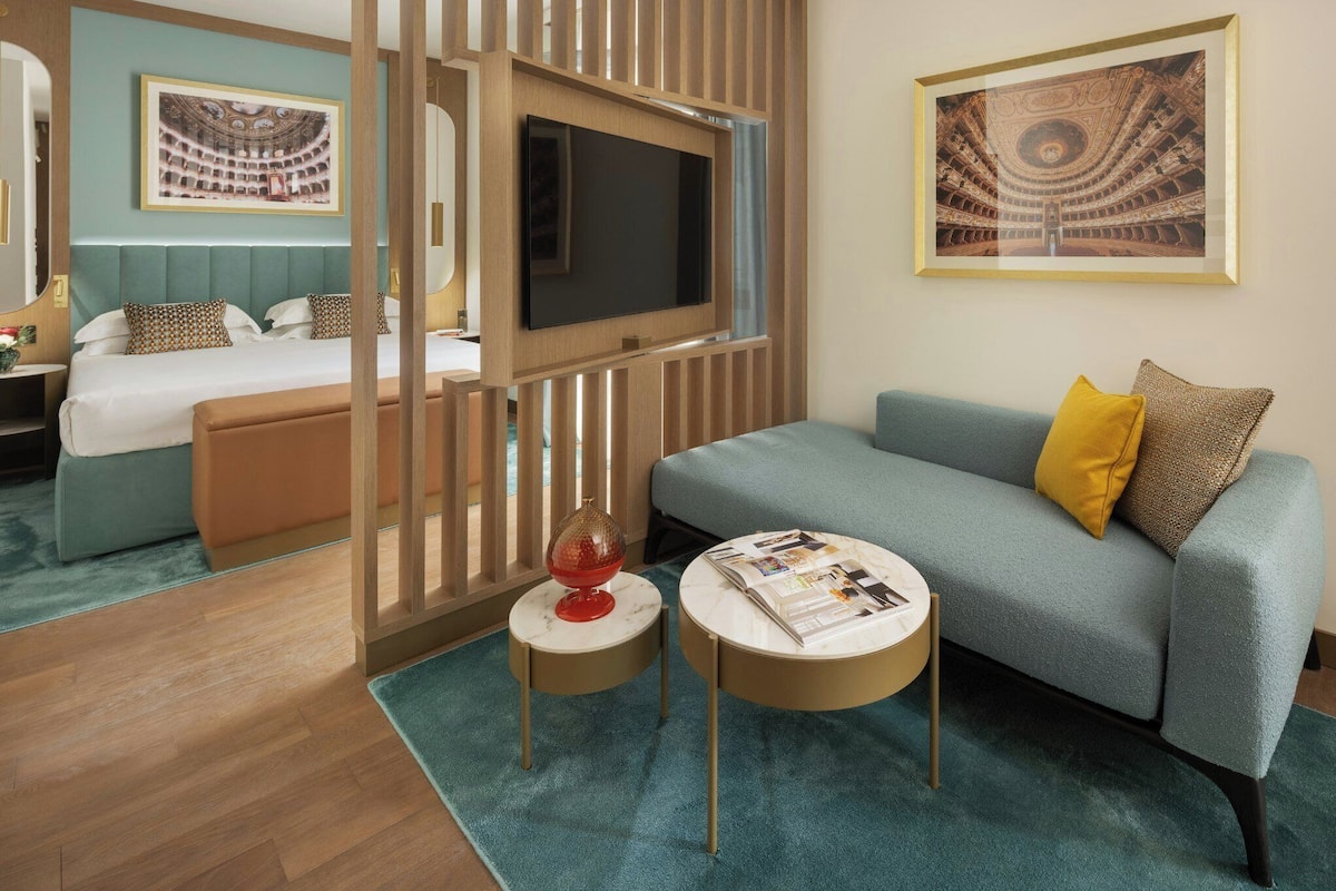 A hotel room with a bed, a TV mounted on a wooden partition, a sofa, two round side tables, and a mirror.