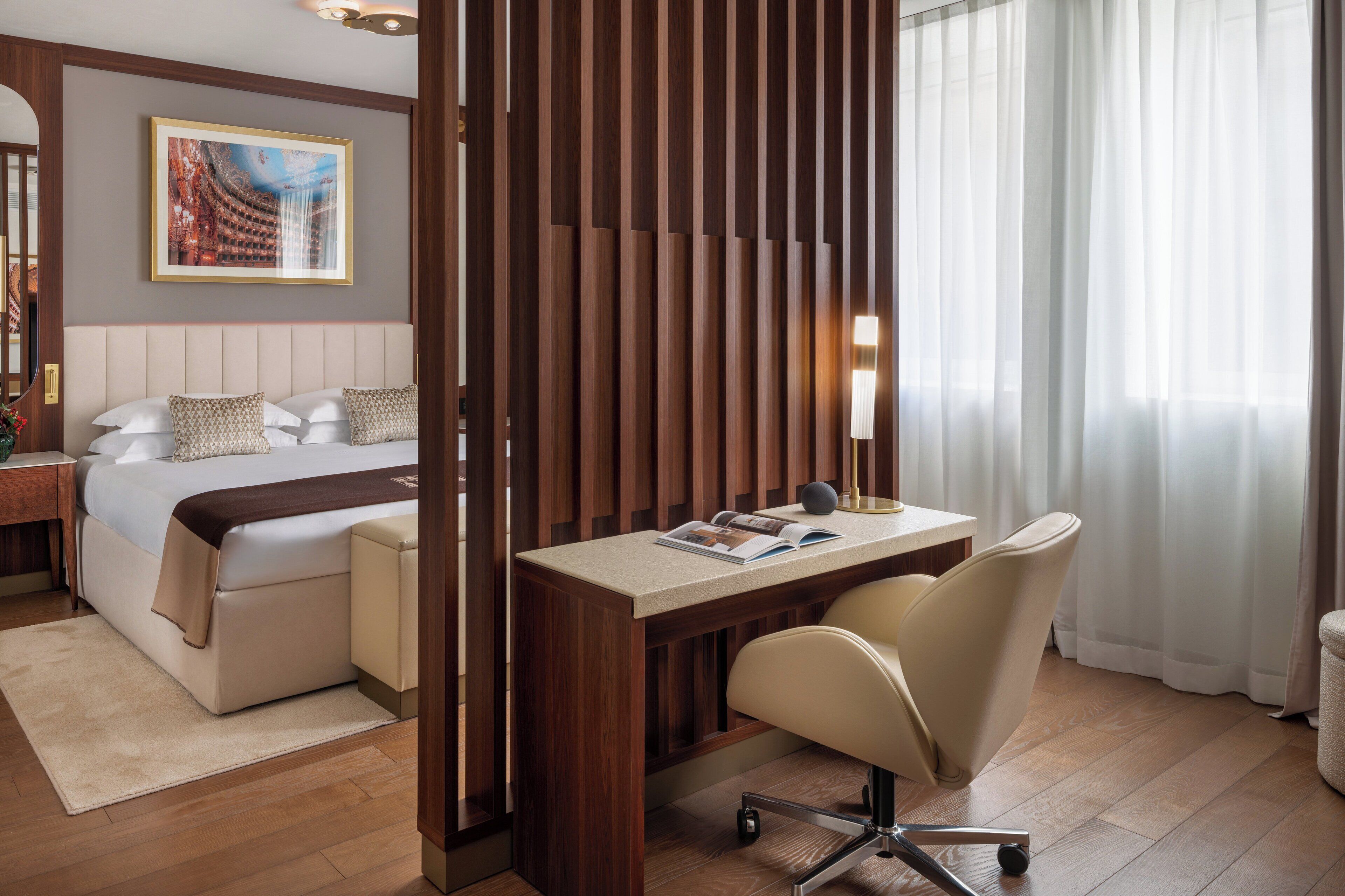 A hotel room with a bed, a desk, a chair, a nightstand, a lamp, and a window with curtains.