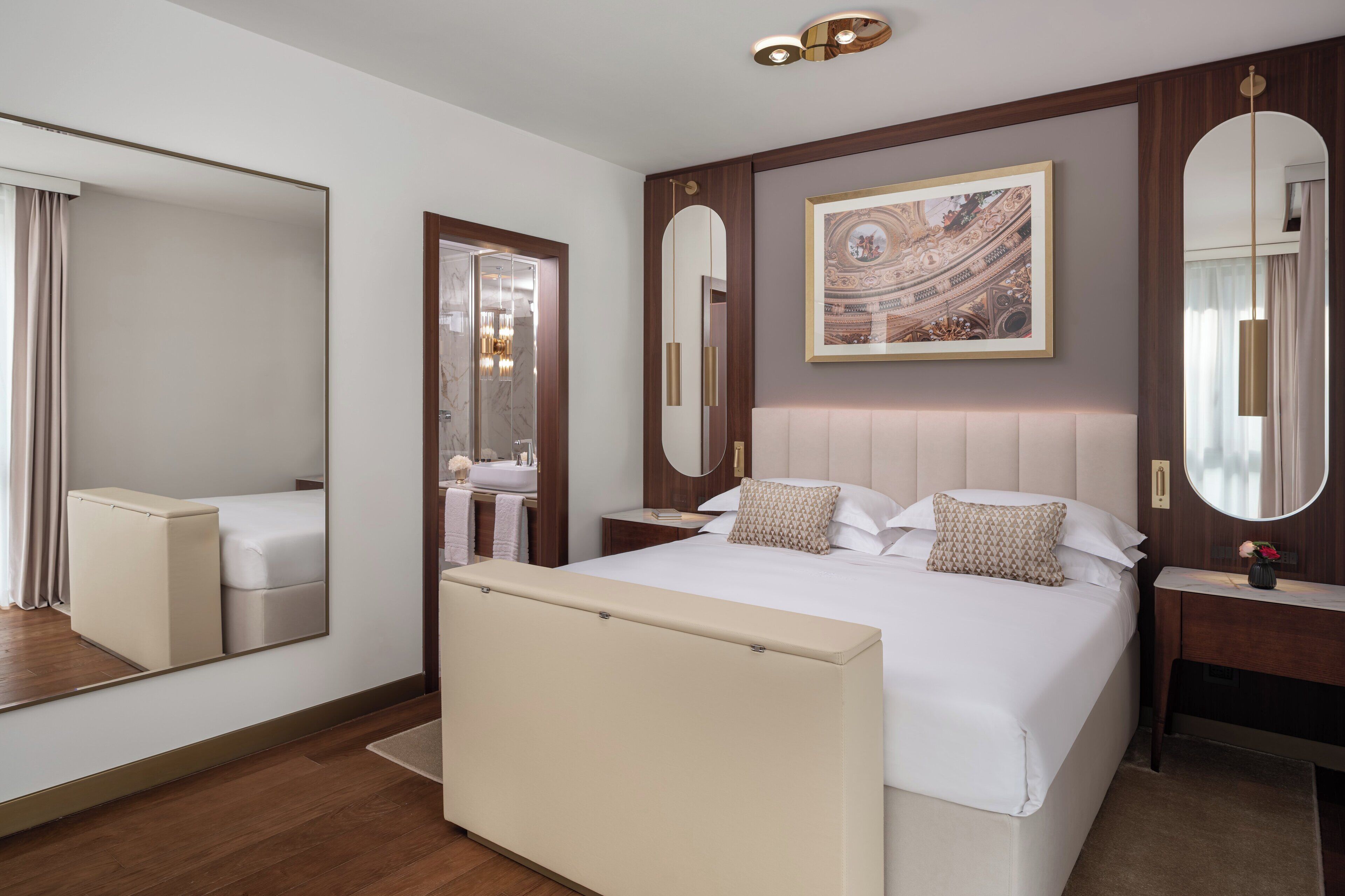 A hotel room with a large bed, a bedside table, a mirror, a wall-mounted artwork, and a large mirror on the wall.
