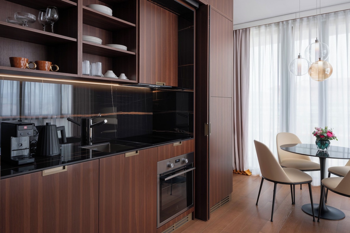 A modern kitchen with dark wood cabinets, a black countertop, and stainless steel appliances. There is a dining area with a round table and chairs, and a window with sheer curtains.