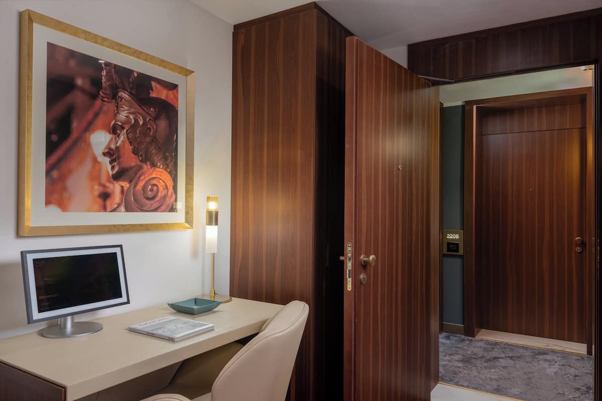 A hotel room with a wooden wardrobe, a desk with a computer, a framed artwork, and a lamp.