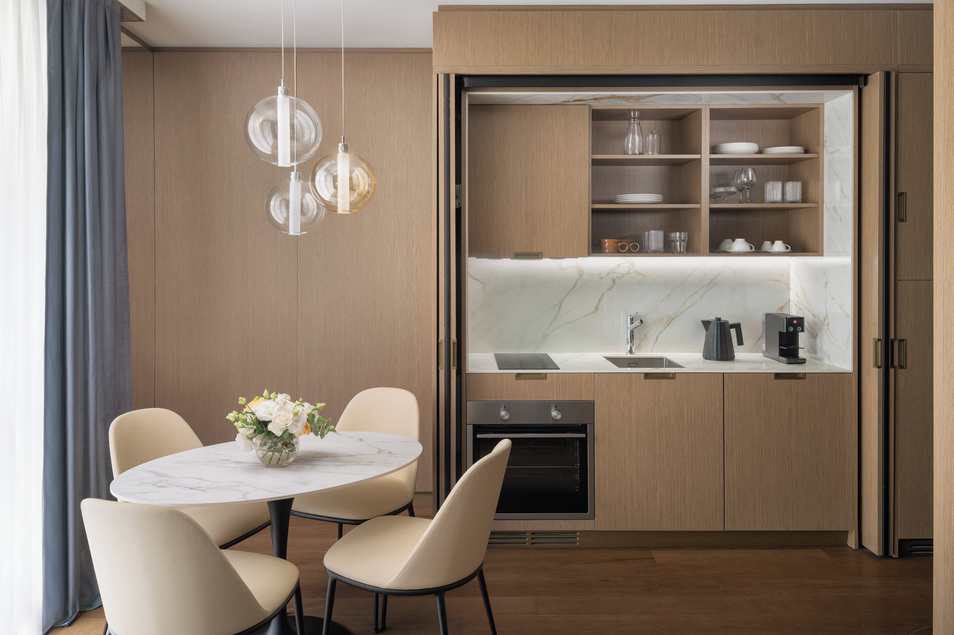 A modern kitchen with a built-in oven, a coffee maker, and a marble countertop. There is a dining area with a round table and chairs, and a vase with flowers.