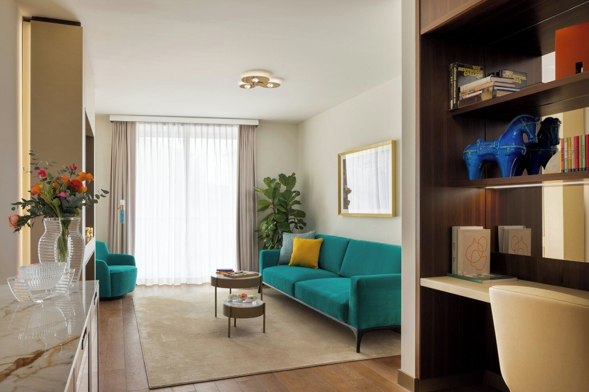 Ein modernes Wohnzimmer mit einem Türkis-farbenen Sofa, einem Couchtisch, einem Bücherregal und einem großen Fenster mit durchsichtigen Vorhängen.