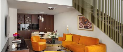 A modern living room with a sofa, armchairs, and a coffee table. There is a kitchen in the background and a staircase with a railing.