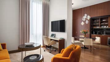 A modern hotel room with a sofa, a desk with a laptop, a dining area, and a TV mounted on the wall.