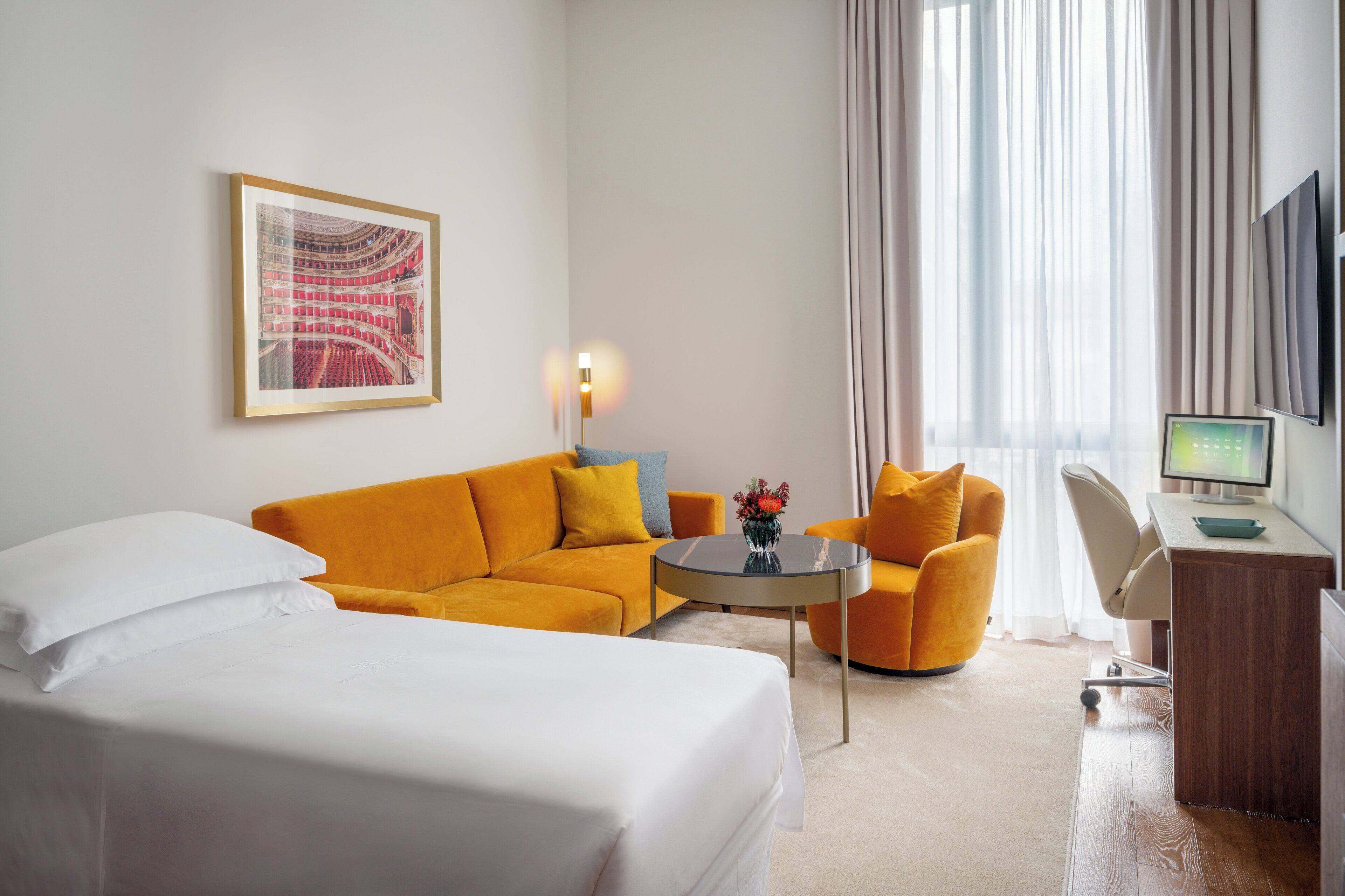 Una habitación de hotel con cama, sofá, escritorio con ordenador y un amplio ventanal con cortinas.