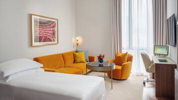 Una habitación de hotel con cama, sofá, escritorio con ordenador y un amplio ventanal con cortinas.