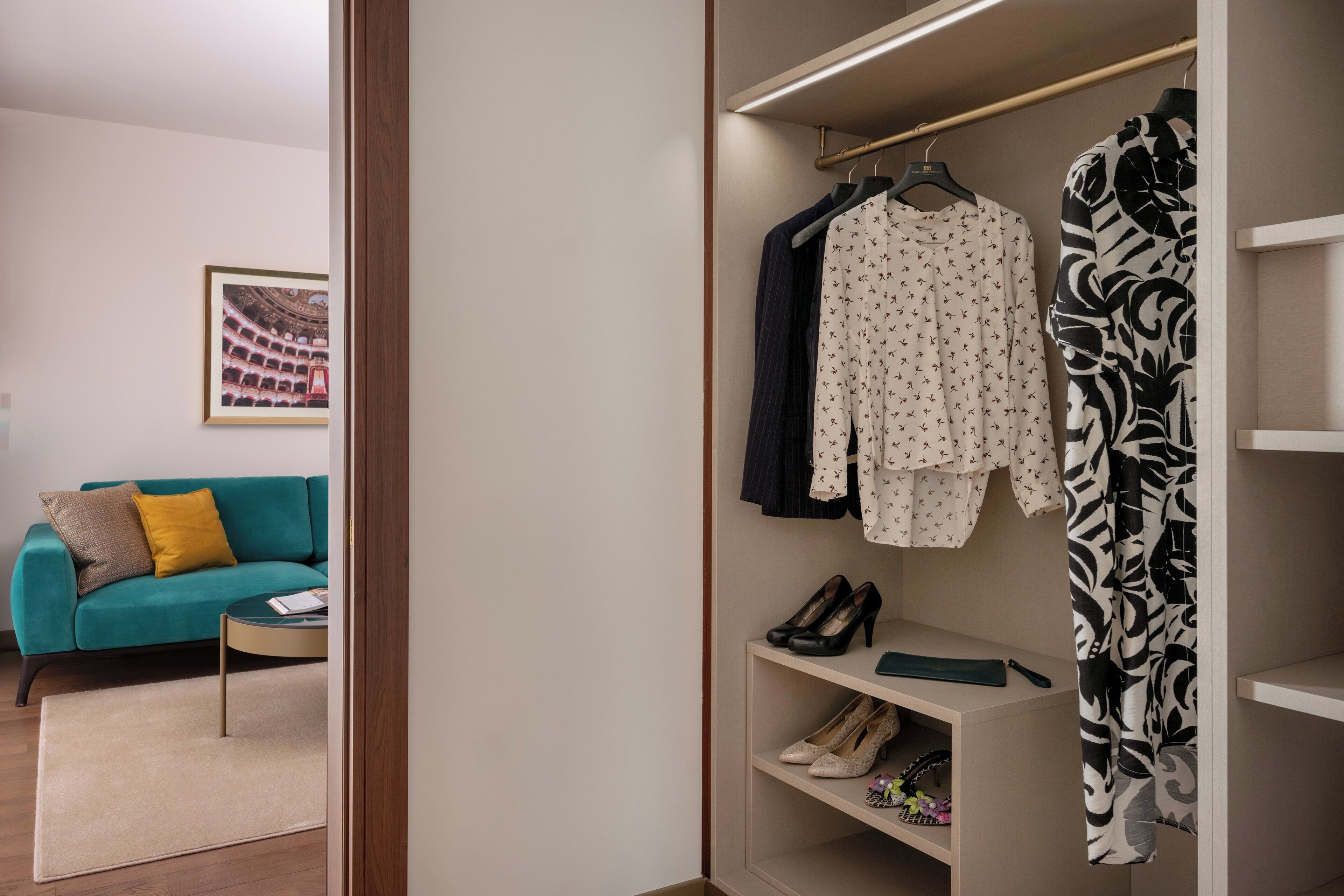 A modern room with a teal sofa, a framed picture on the wall, and a closet with hanging clothes and shoes.