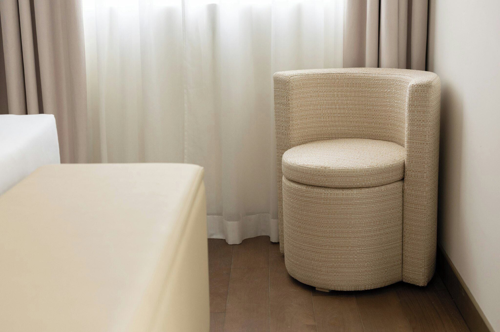 A corner room with a single bed, a woven chair, and a window with sheer curtains.