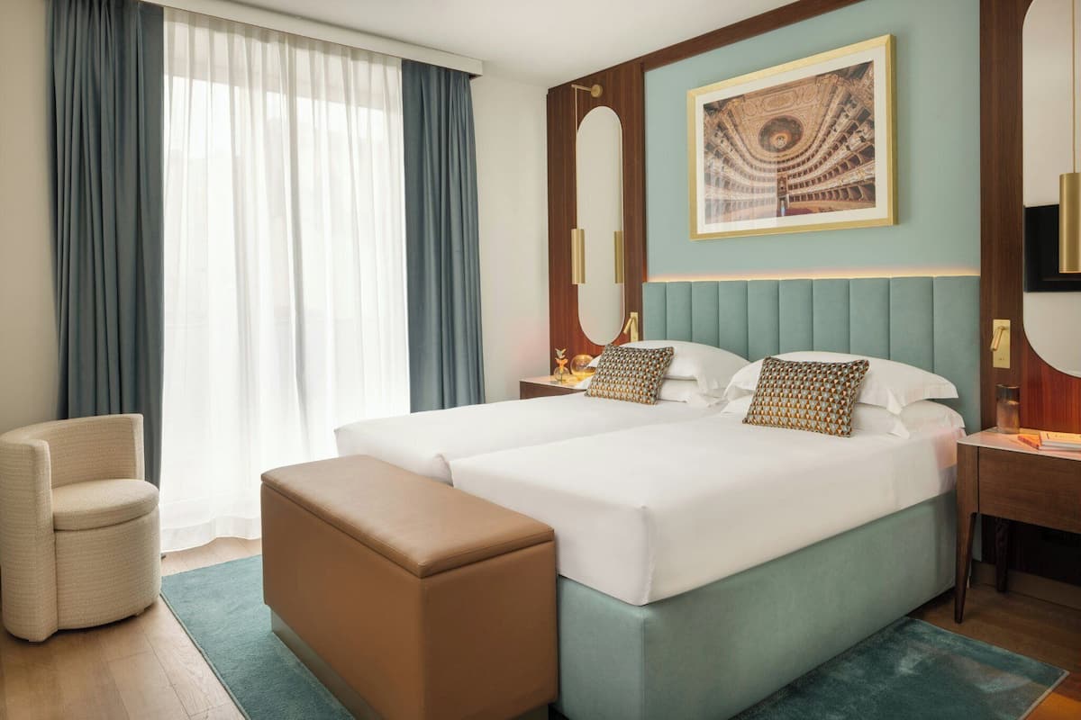 Habitación de hotel con dos camas, una zona de estar, un ventanal grande con cortinas, un cuadro enmarcado en la pared y una mesita con lámpara.