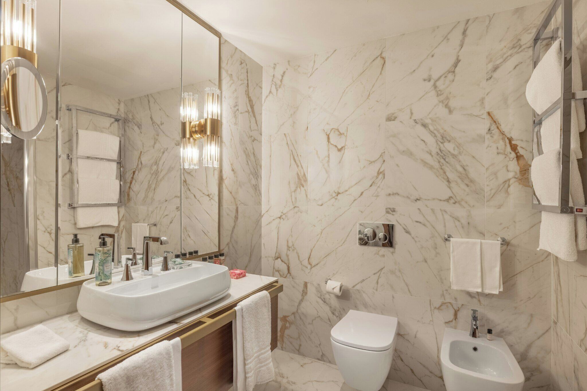 Un baño moderno con un espejo grande, paredes de mármol, un lavamanos blanco y un bidé.