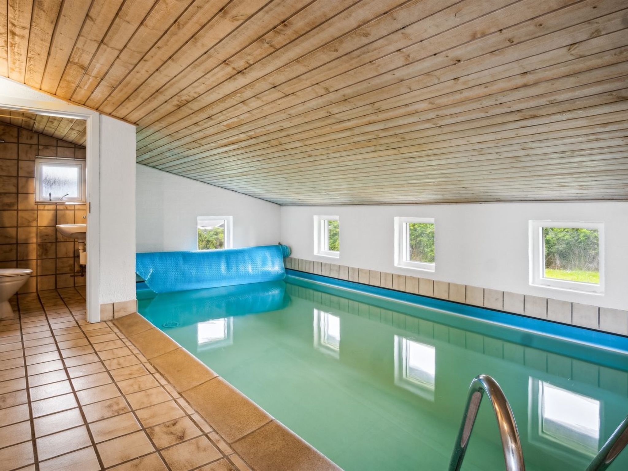 An indoor swimming pool with a wooden ceiling, a blue bench, and small windows.