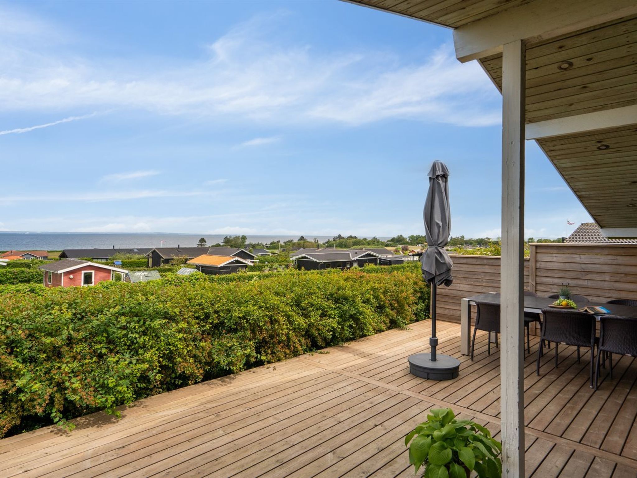 Um deck de madeira com mesa e cadeiras, um guarda-sol e vista para as casas e o mar.